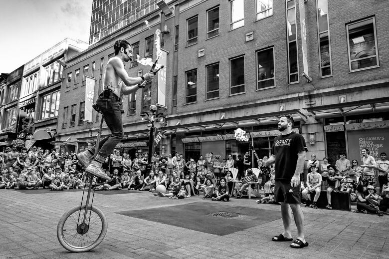 A shirtless man on a unicycle juggles three fire sticks while another man holds another stick from the ground.