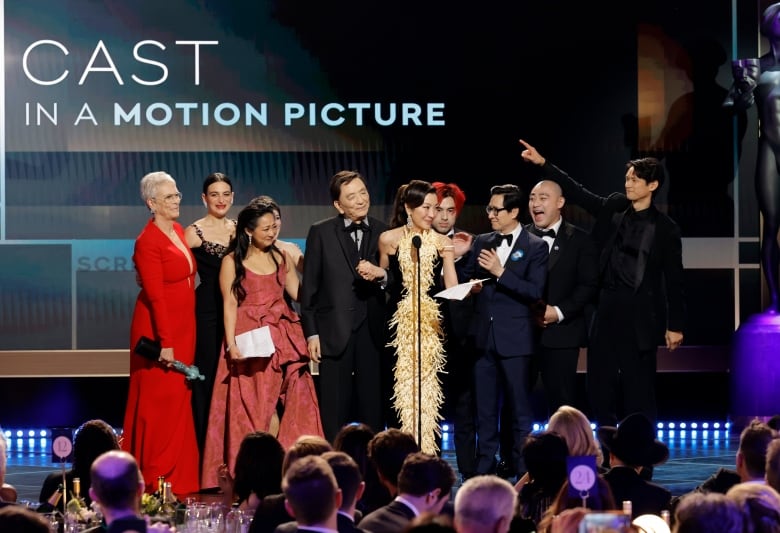 A group of ten stand onstage celebrating. Behind them the words 