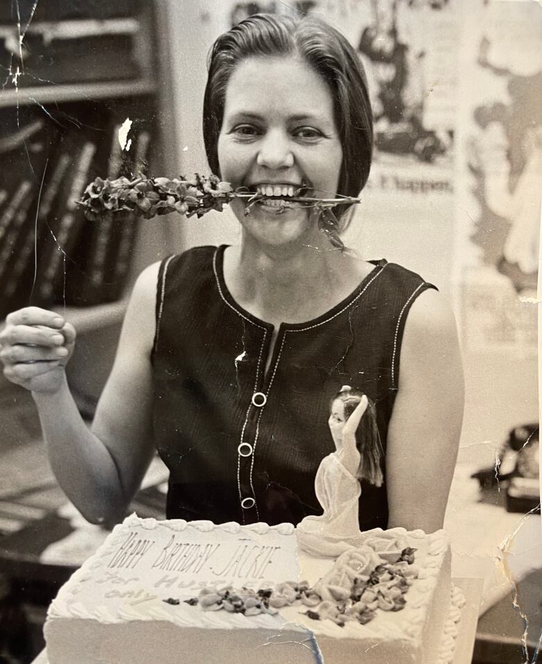 A black and white photo of a smiling woman with a flower in her mouth, holding a birthday cake.