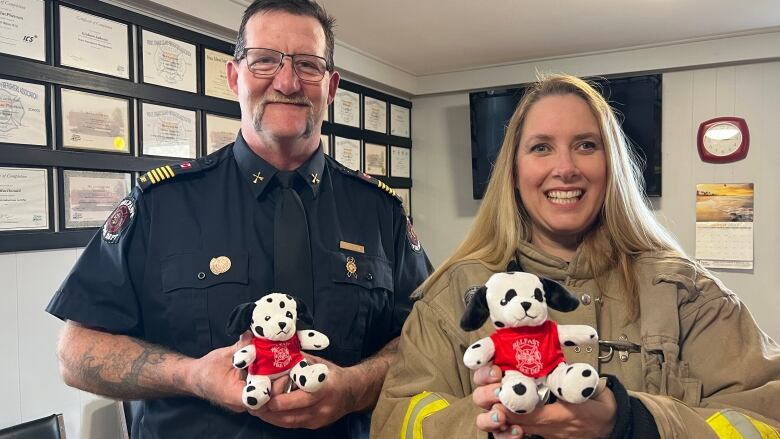 A man and a woman in firefighter gear, smiling for the camera. Both are holding stuffed toy Dalmatian dogs, which have a red shirt with a logo.