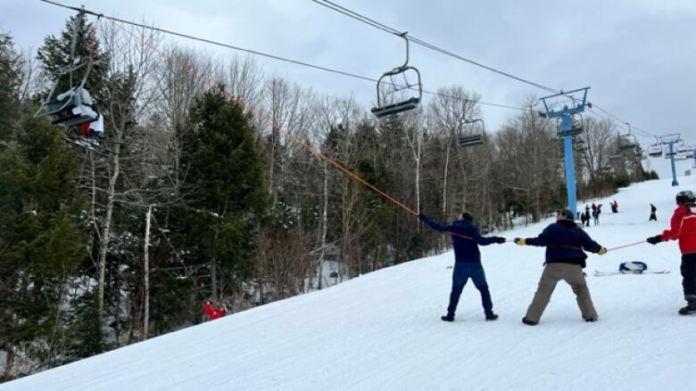 A photo of a ski hill's chairlift taken from the ground. A team of five people to the right of the photo hold one end of an orange rope that extends over the chairlift cables, and another person on the far left holds another rope as they all look up.