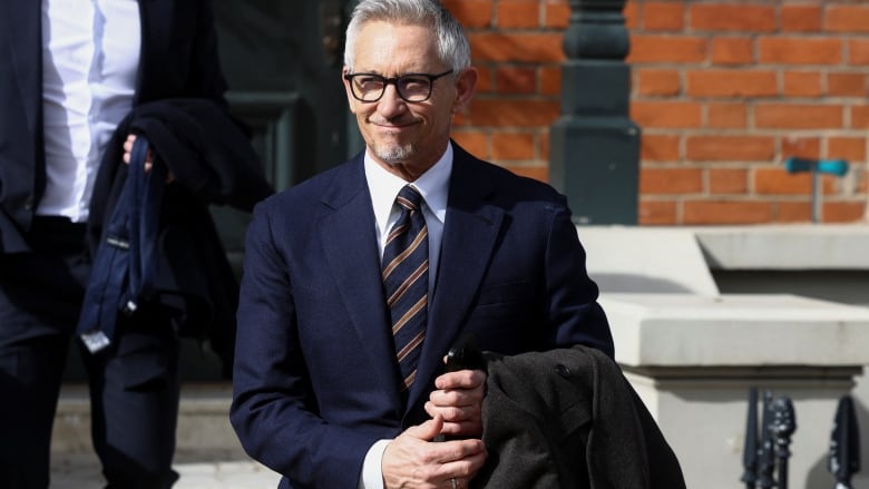 A man wearing styled grey hair, wearing glasses and suit and tie, exits a brick building. 