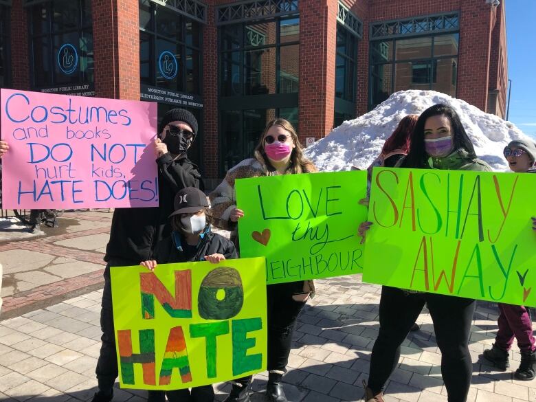 Three adults wearing medical masks stand in a light holding posterboard signs. The first is pink with 