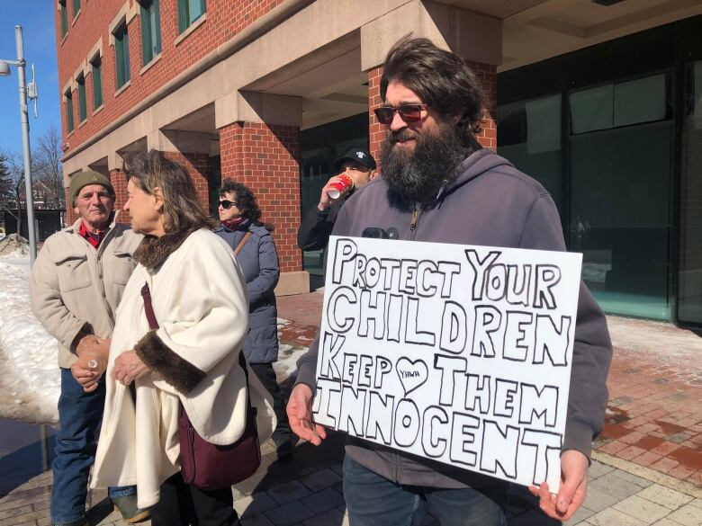 A man in a beige coat and jeans stands to the left of the photo, with his hands held together in front of him, a woman with short dark hair and a light beige coat stands next to him, looking into the distance to her right, and a man with long dark hair and a long dark beard stands closest to the camera, holding a white sign that says 