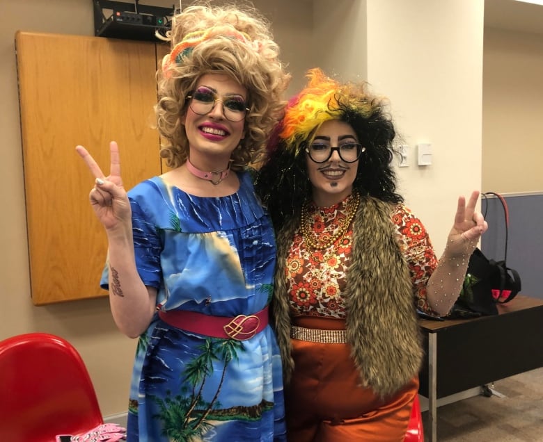 A drag queen with big blonde curly hair stands on the left, smiling at the camera and holding up two fingers. She is wearing a blue dress depicting a beach scene, with a red belt at her waist, and glasses. She has her arm around another drag queen, who has mostly curly black hair with bright yellow and orange highlights on top. She is also wearing glasses, a colourful orange and creme floral long sleeve shirt with a brown faux fur vest. She has a dark orange skirt and gold belt, and is also holding two fingers up to the camera in 