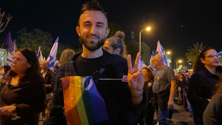 Smiling man with beard holding a rainbow flag and fingers in a peace sign on a crowded street at night.