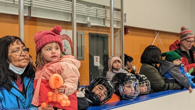 The photo shows several people standing behind the ice rink, including three small children wearing hockey gear. 