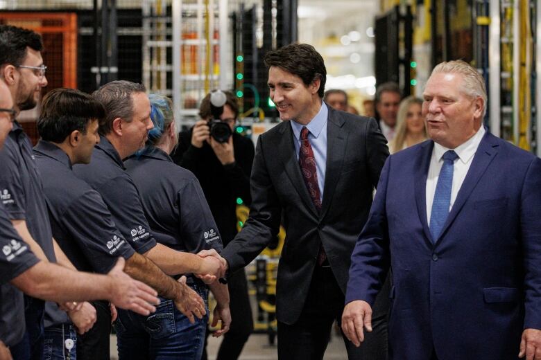 Two male government leaders in suits meet with electric vehicle manufacturing plant workers during a grand opening.