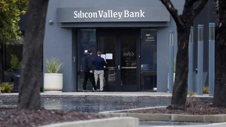 A man puts a sign on a door of a building with a Silicon Valley Bank sign as another man watches. 