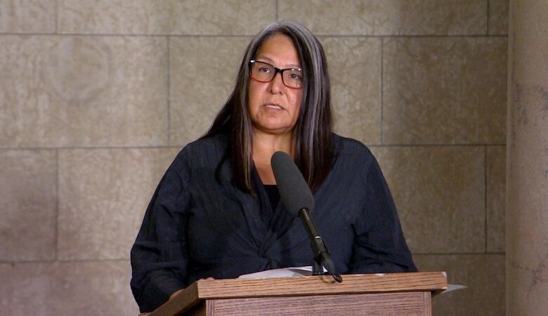 A woman with long black hair, streaked in white, wears glasses and speaks into a microphone.