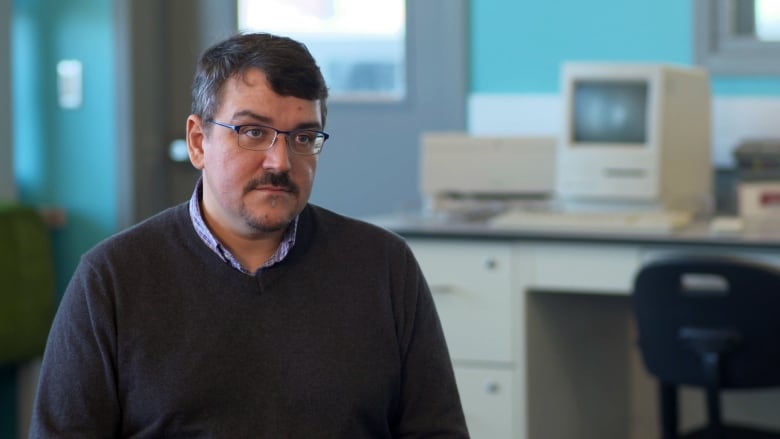 A man in a navy sweater with greying brown and half-rimmed glasses. He's sitting in a computer lab.