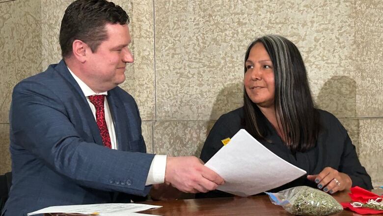 A man in a suit and red tie sits at left holding a piece of paper and smiling at a woman to his left. She smiles back and has long black hair with white streaks in it.