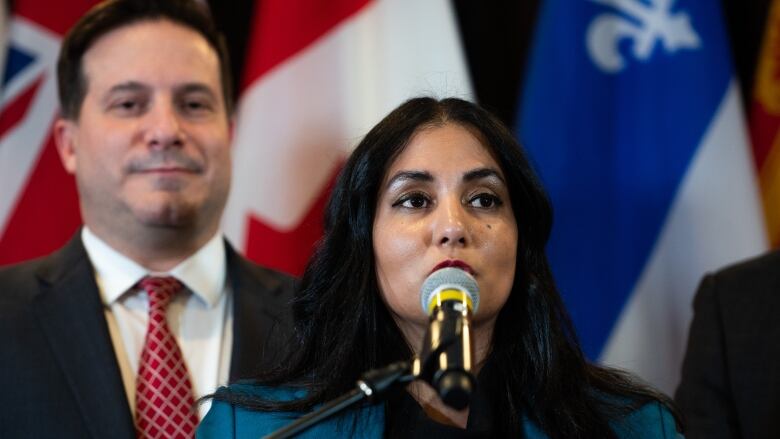 A woman with shoulder-length black hair and a blue blazer speaks during a news conference. The flags of Canada and Quebec are behind her.