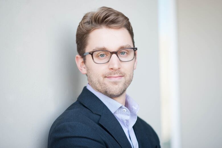 A man with brown hair, stubble and glasses leans against a wall in a blue shirt and jacket. 