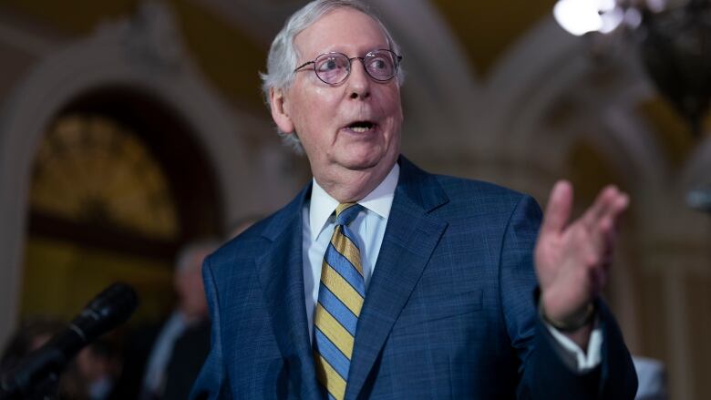 White-haired man with glasses wearing a blue suit and yellow and blue striped tie, speaking with one hand up