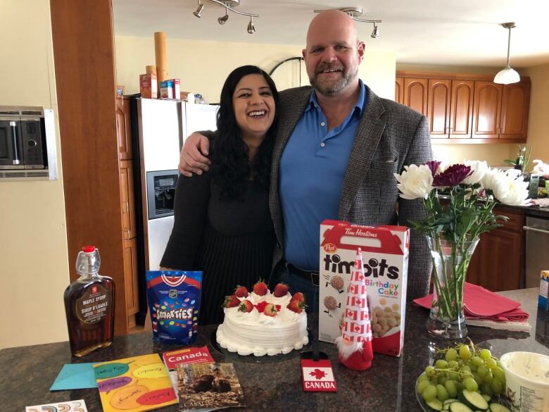 Women with brown hair and dark shirt stands beside man in brown blazer. The stand behind a kitchen counter with maple syrup bottle, cake and cereal box.