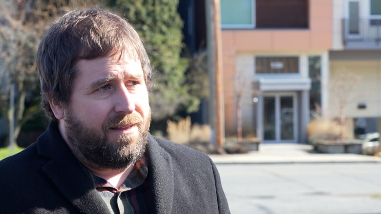 A young man with a beard in an overcoat stands in front of an apartment building.