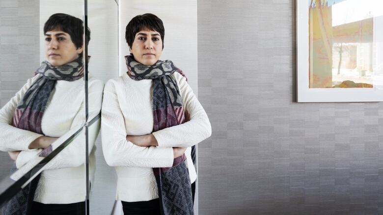 A woman with short, dark hair wearing a scarf stands against a wall and looks at the camera with a solemn expression on her face.