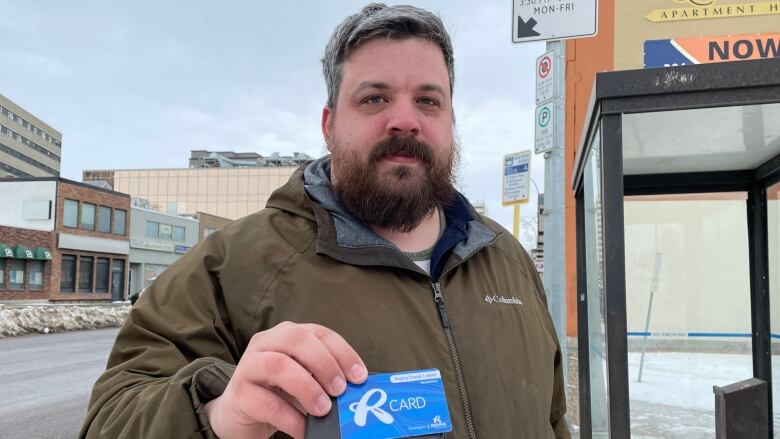 A bearded man wearing a brown Columbia jacket holds his wallet and an R-Card. 