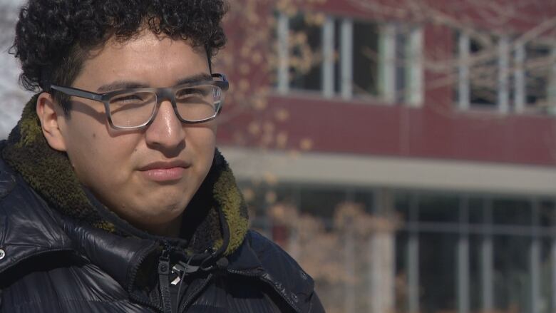 A young man, in a black coat and scarf standing outside a building at the University of Winnipeg campus. 