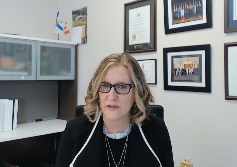 A woman with blond hair and glasses sits at a desk with framed photos on the wall behind here.