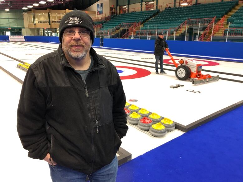 A man with a beard and black jacket standing in front of some curling rinks.