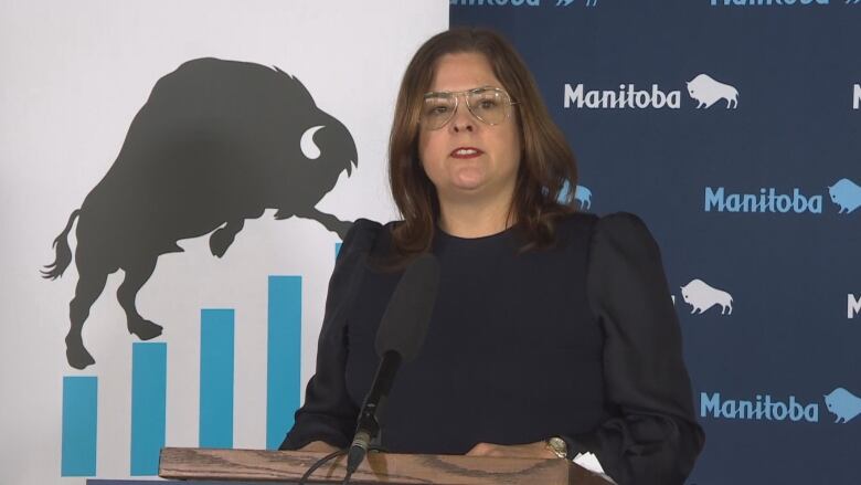 A woman with long brown hair and glasses speaks at a podium. Behind her, on a backdrop, is the word Manitoba repeated over and over, with a silhouette of a bison. 