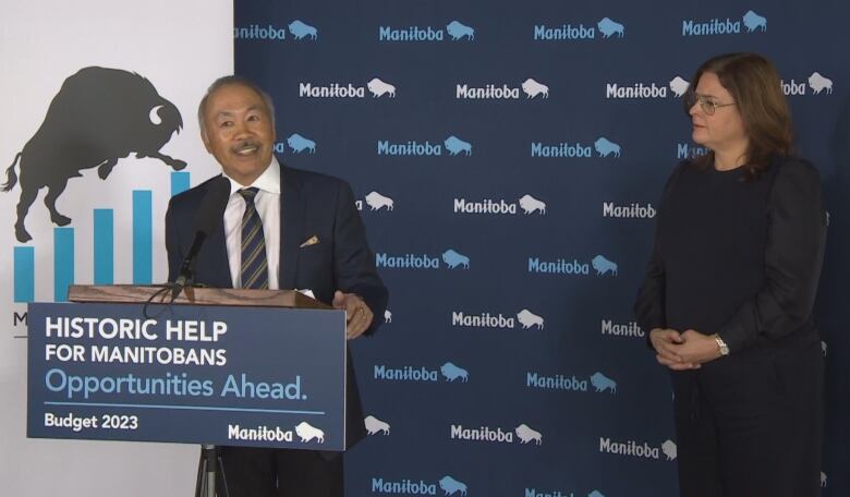 A balding man with a moustache and wearing a suit smiles as he speaks from behind a podium. A sign on the podium says 