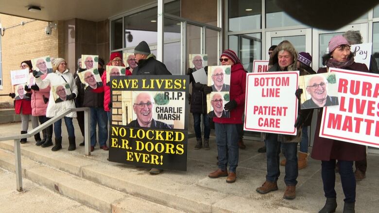 Several people stand on the steps of confederation building holding posters. The posters read statements such as 