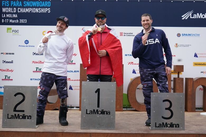 Three men show off podiums at a sporting event. The person in the centre has a red and white flag draped around them.