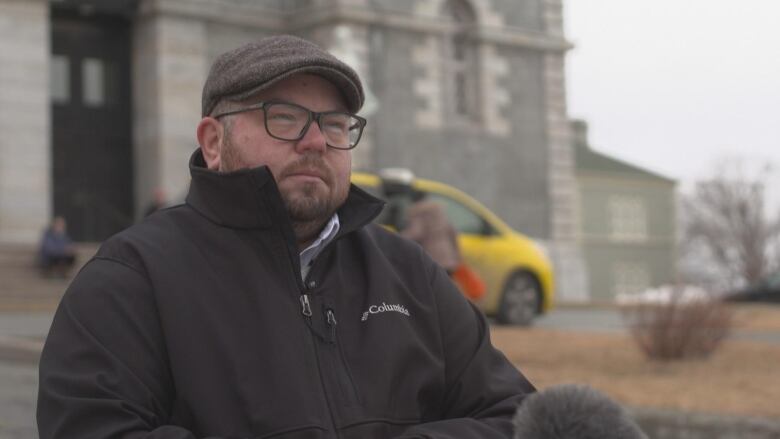 A person wearing a grey wool cap and black jacket, with dark-rimmed glasses and a short beard.