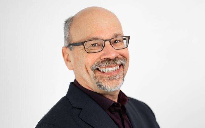 A man with grey hair and a trim beard smiles at the camera in a professional pose. He is wearing glasses and a dark blazer.