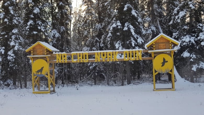 A large sign in front of a snowy forest reads 'Na-Cho Nyk Dun.'