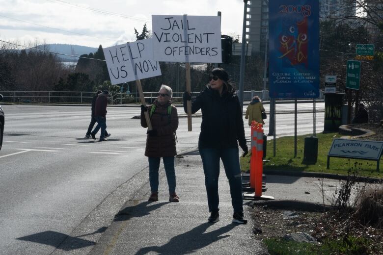 Two women are holding placards that read 'jail violent offenders' and 'help us now' on the sidewalk.