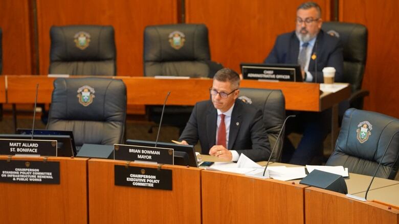 A man in a suit sits behind a wood-panelled row of desks in the city council chamber.