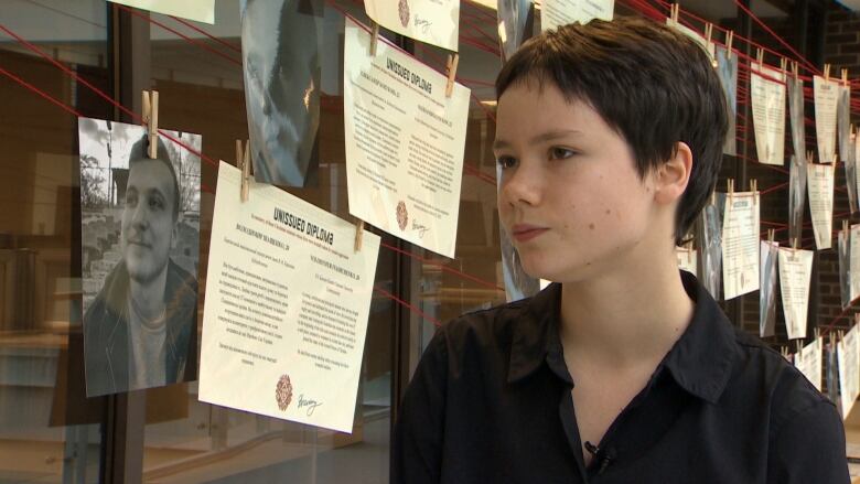 Kateryna has short brown hair and a black collared shirt. She is standing next to the photos of students in the exhibit, which are held up by paper clips across red thread.