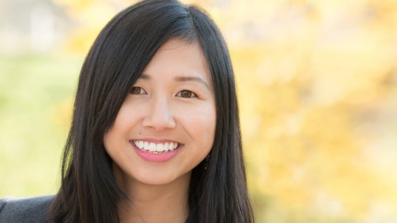 A woman with straight brown hair is wearing a black blazer with a bright pink blouse and is smiling at the camera. She's standing outside in a park which is blurred. 