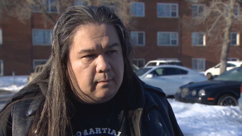 A man with long dark hair stands in front of an apartment building. 