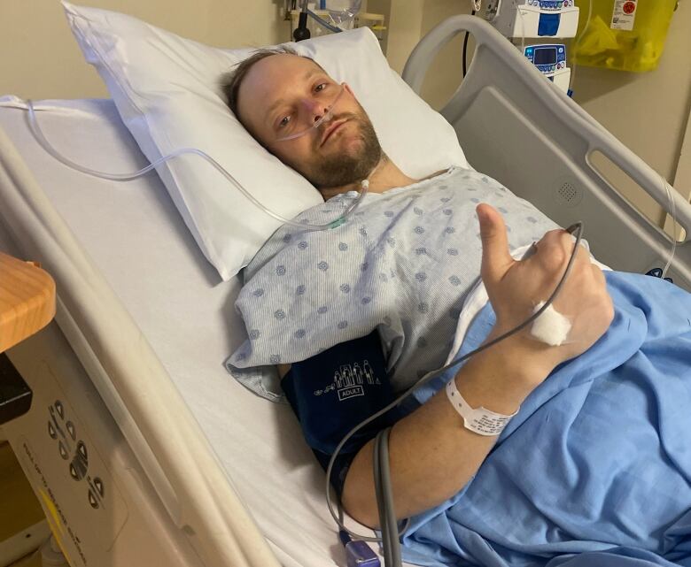 A man lying in a hospital bed wearing a blue hospital gown. He has oxygen tubing in his nose and a blood pressure cup around his arm.