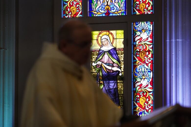 A priest speaks during a church service. The priest is blurry, and a stained glass window is in focus. 