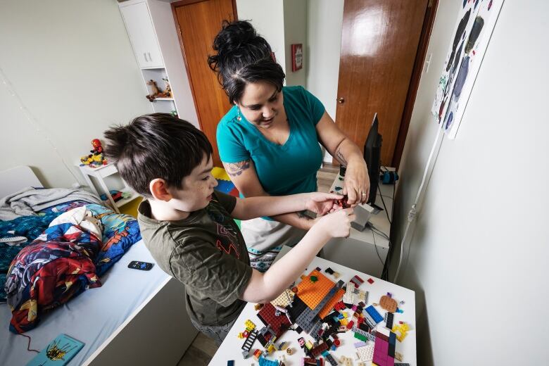 A young boy stands at a desk in his room with his lego, showing his mom something he built. 