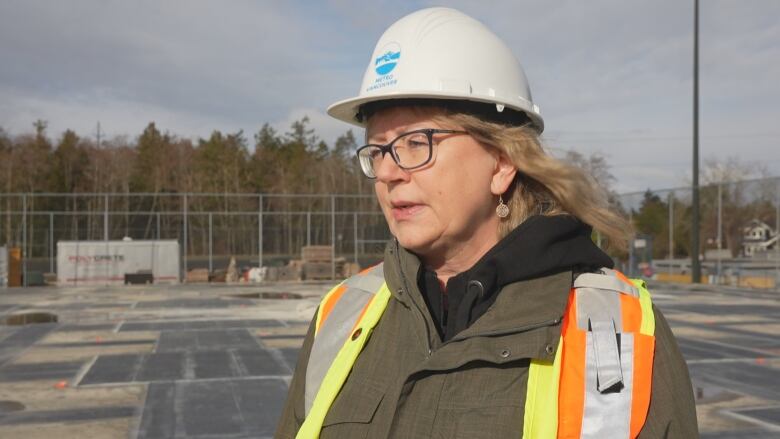 A woman with short blonde hair stands on a concrete ground, wearing a hard hat and bright yellow and orange vest.