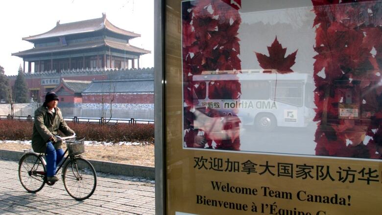 Man on bicycle in front of Chinese building, with Canadian flag on billboard.