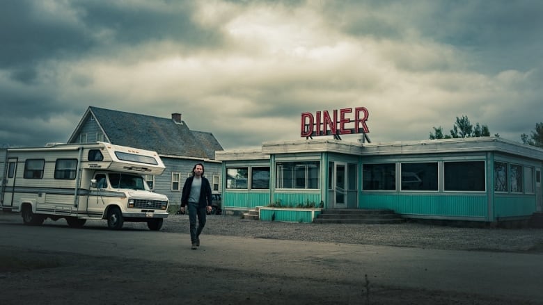 An actor stands in front of a diner on a film set. The sky is cloudy.