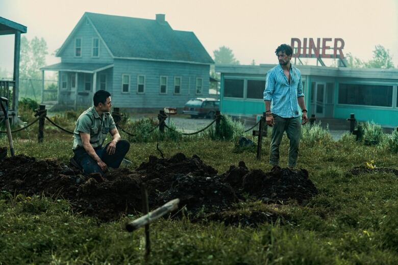 Two men stand in a makeshift graveyard, looking at each other. There's an old house and diner in the background.