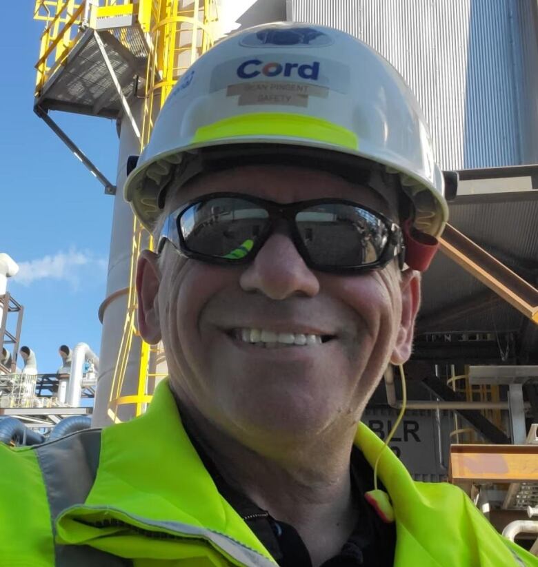 A middle-aged man wearing a hard hat and sunglasses smiles.