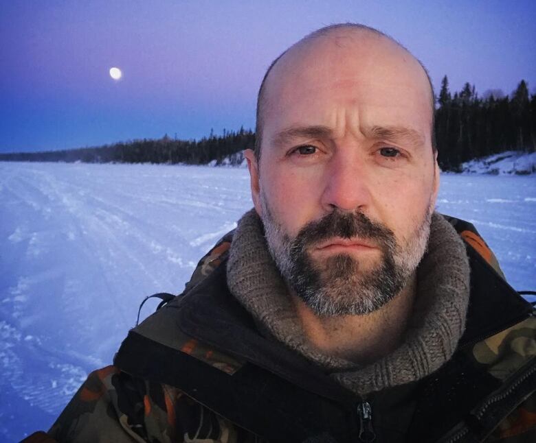 A man stands on a lake covered in snow, with the moon in a dusky sky behind him.