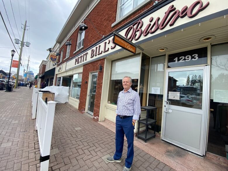 A man stands in front of a restaurant. The words Petit Bills Bistro are on the storefront sign above him. 