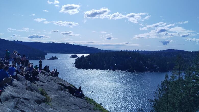 A number of people gather on a large rock to the left of the image, with a panoramic view of islands, hills and the sea visible.