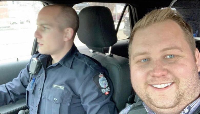 A man smiles as he sits besides a police officer in a car. 